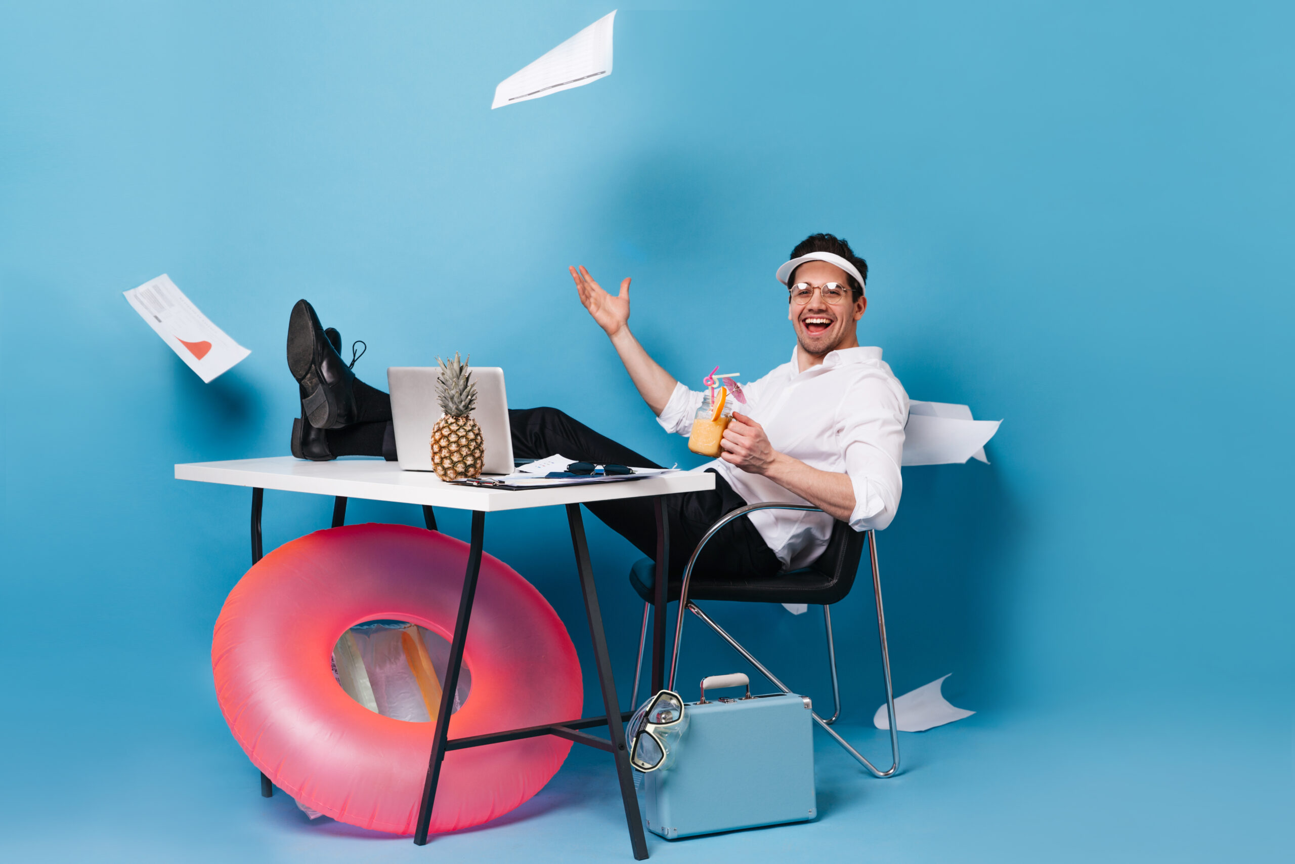 Perky businessman in suit and cap scatters graphics, enjoying tasty cocktail, sitting at table with laptop, rubber ring, suitcase on blue background